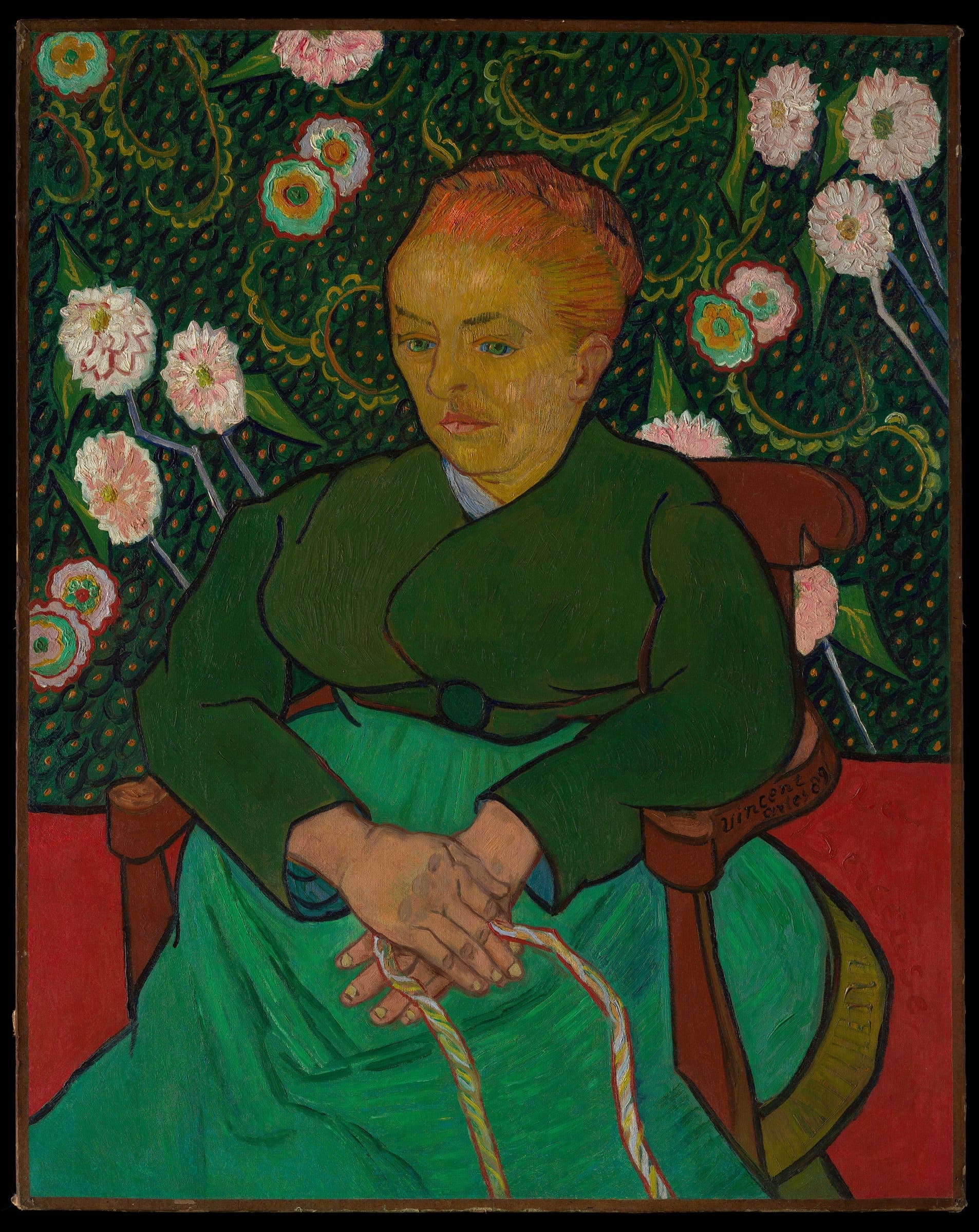 A seated woman in a green jacket holds a cord, against a patterned wall of flowers in red, green and orange.